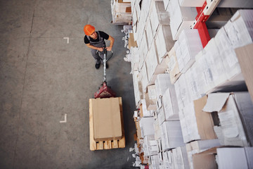 Top view of young male worker in uniform that is in the warehouse pushing pallet truck