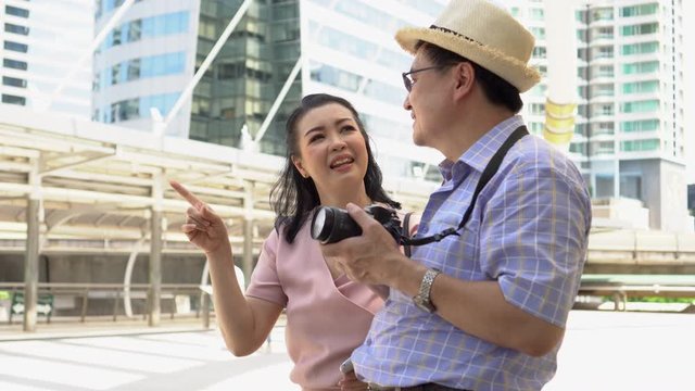 happy asian Senior couple tourists traveling with camera Pointing finger to destination in urban city outdoors. old man Travellers lifestyle  . elderly woman vacation . ageing society concept.