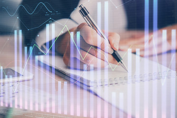A woman hands writing information about stock market in notepad. Forex chart holograms in front....