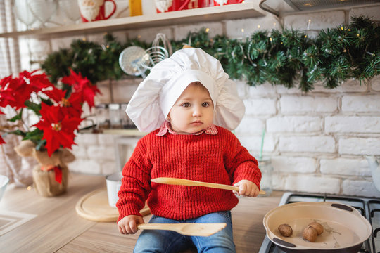 one year old girl in a cook hat and with kitchen utensils sits in the kitchen decorated for Christmas