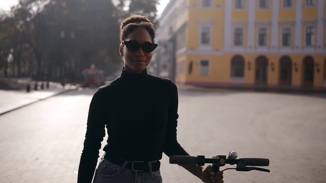 Attractive, confident afro american girl in stylish sunglasses and blue jeans walking carefree by morning city street with her black electric scooter. Front view. Slow motion