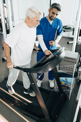 Elderly man doing cardio workout on a treadmill, therapist helping senior patient to recovery