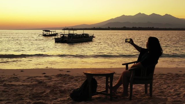 Girl sitting on a chair over fine sand of empty beach photographs silhouette of boats floating on calm lagoon few minutes after sunset in Thailand