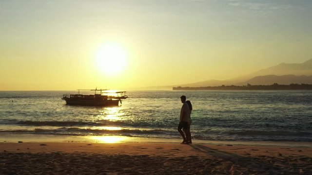 Silhouette of young couple walking on calm beach at romantic sunset moments alongside boats floating on sea of Malaysia