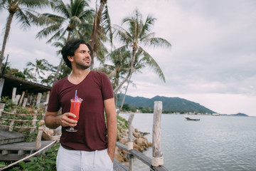 A young man stands by the sea on a wooden bridge with a cocktail.
