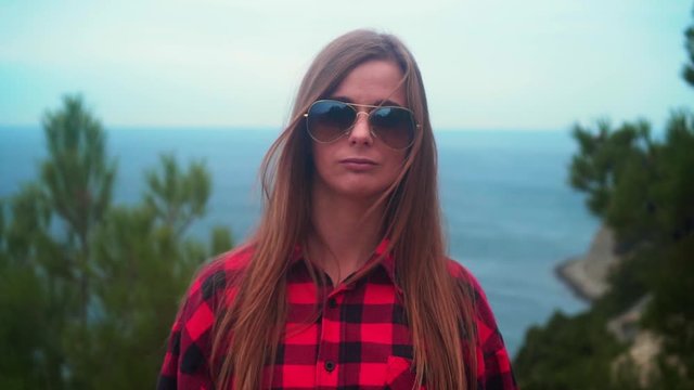 Portrait of a sexy girl with long hair in a black and red shirt and sunglasses, stands against the backdrop of the sea. Against the background of the blue sea.