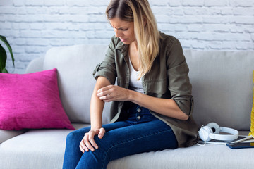 Uncomfortable young woman scratching her arm while sitting on the sofa at home.