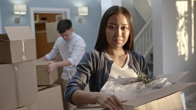 Tilt up of happy Asian woman holding cardboard box and packing potted plant with bubble wrap, then looking at camera; young man putting boxes with belongings in pile in background