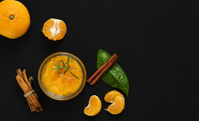 Citrus (tangerine) jam in a glass jar on a table with a cinnamon stick and a branch of rosemary. Nearby are slices of mandarin, a leaf of mandarin and a whole fruit. Top view, copy space. 