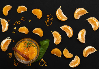 Citrus (tangerine) jam in a glass jar on a table with a cinnamon stick and a branch of rosemary. Around segments of mandarin, shavings of zest. Top view. Black stone background.