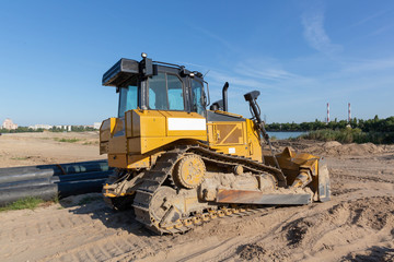 A crawler, yellow, black-roofed bulldozer stands on the sandy surface of the dam under a blue sky.