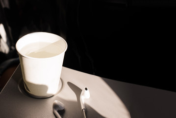 White paper cup of water and headphones on a folding table in an airplane. Drinks on the plane. Dehydration in flight. skin hydration