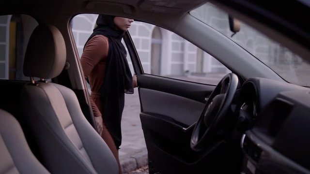 Young beautiful muslim girl in black hijab open the door and sitting in car, watching at mirror, fixing it. Insert the keys - starts the engine, putting on the safe belt - ready to drive. Side footage