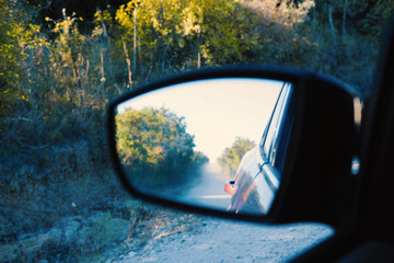 Rural dirt road reflection in car rear view mirror, driving through scenic countryside concept.