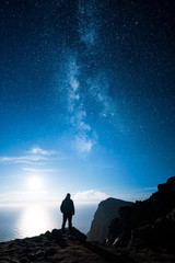 A lonely man standing by a cliff and looking at the ocean and stars of the milky way