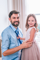 daughter tying tie on her father and smiling at the camera in living room