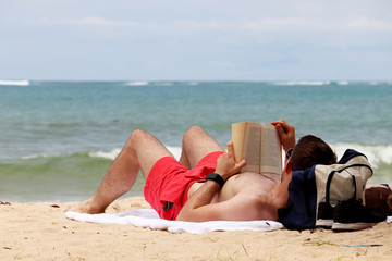 Man lying on the sand, sunbathing and reading a paper book. Beach leisure on a seacoast, relax, reading guy