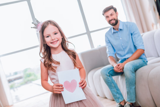 girl holding picture of heart and looking at the camera while father sitting on the sofa at home
