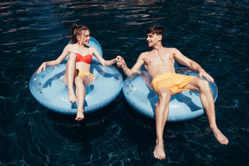 happy young couple holding hands while relaxing on swim rings in swimming pool