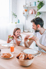 father and smiling daughter eating sandwiches for breakfast on the kitchen