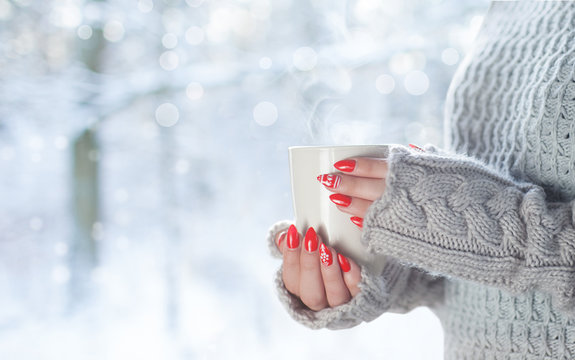 Young woman with morning cup of coffee looking through the window, blurry winter snowy woods outside.