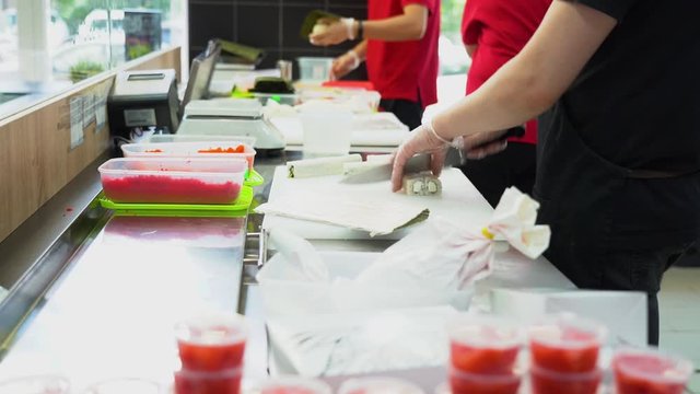 Asian people in red t-shirts cooking rolls. Sushi master in gloves skillfully cuts rolls with a sharp knife on a white table in the big kitchen. Rolls with rice and fish prepare for sale on dinner.