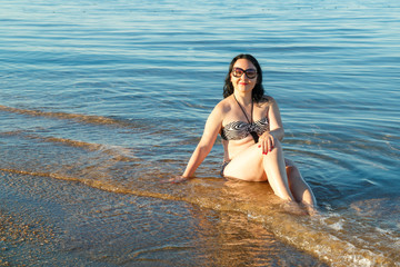 Fototapeta na wymiar A brunette woman in a swimsuit and glasses sits on the seashore.