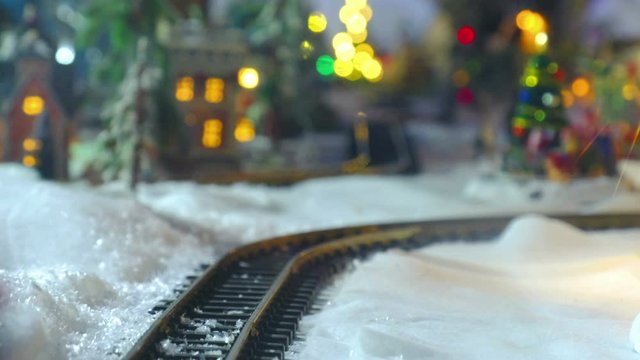 Christmas Toy Train Model Close Up Tracks On The Snow With Town Village Lights On Background