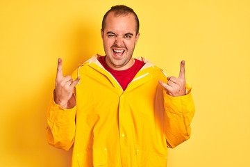 Young man wearing rain coat standing over isolated yellow background shouting with crazy expression doing rock symbol with hands up. Music star. Heavy concept.