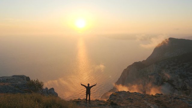 Silhouette of a Young Woman standing on a cliff edge observing a beautiful dramatic sunset over a sea from a high mountain in Crimea. Lady hiker with arms outstretched against beautiful sunset light.