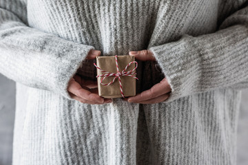 Girl in a knitted winter sweater holds out a small hand made Christmas gift box in craft paper and bow rope. DIY. Concept