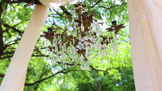 Wedding decor. Beautiful luxury decorations for outside wedding celebration. Closeup of christal chandelier without bulbs hanging in green summer park or wood. Arch decorated with draped fabric.