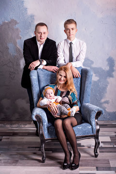 Family Portrait Of Four. Mom And Baby Boy Are Sitting In A Chair While Dad And His Teenage Son Are Standing Behind Them