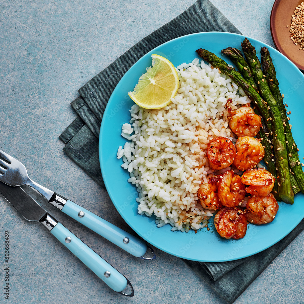 Wall mural Delicious teriyaki coated shrimps stir fry with grilled asparagus, rice and sesame on a blue plate. Healthy seafood meal. Top view, directly above shot.