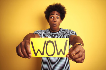 American man with afro hair holding wow banner standing over isolated yellow background scared in...