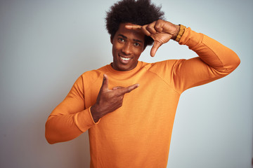 Young american man with afro hair wearing orange sweater over isolated white background smiling making frame with hands and fingers with happy face. Creativity and photography concept.