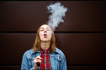 Vaping teenager. Young pretty white girl in casual clothes smoking an electronic cigarette opposite dark modern background on the street in the autumn. Bad habit. Vape activity.