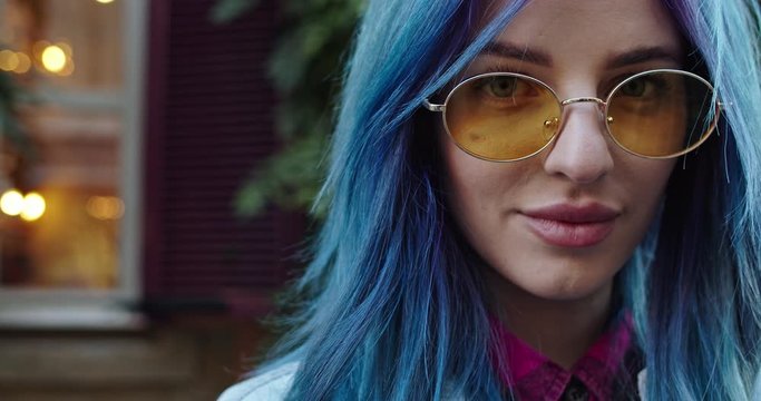 Portrait of the face of the young beautiful and charming Caucasian hipster girl with blue hair and in sunglasses rising eyes, looking straight to the camera and smiling happily. Outdoors. Close up.