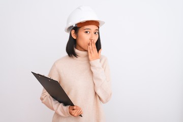 Chinese architect woman wearing helmet holding clipboard over isolated white background cover mouth with hand shocked with shame for mistake, expression of fear, scared in silence, secret concept