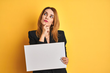 Redhead business caucasian woman holding banner over yellow isolated background serious face thinking about question, very confused idea