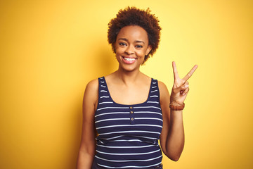 Beauitul african american woman wearing summer t-shirt over isolated yellow background smiling with happy face winking at the camera doing victory sign with fingers. Number two.