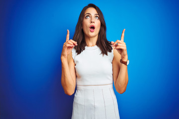 Young business woman wearing white elegant dress standing over blue isolated background amazed and surprised looking up and pointing with fingers and raised arms.