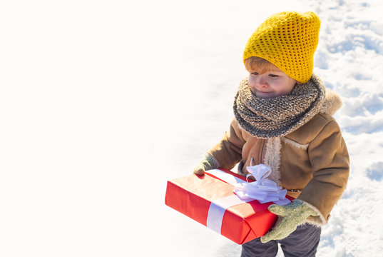 Kid in winter clothes holds Christmas gift. Kid during stroll in a snowy winter park. Beautiful winter nature. Amazing winter park. Adorable kid with cute face. Ptoto with empty copy space.