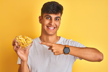 Young indian man holding bowl with dry pasta standing over isolated yellow background very happy pointing with hand and finger