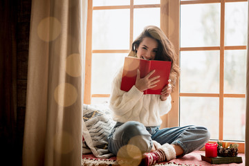 brunette woman in warm sweater and socks reading book sitting on windowsill in room decorated for...