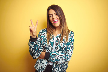 Young beautiful woman wearing casual jacket over yellow isolated background smiling with happy face winking at the camera doing victory sign with fingers. Number two.