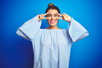Young beautiful woman wearing bun hairstyle over blue isolated background Doing peace symbol with fingers over face, smiling cheerful showing victory
