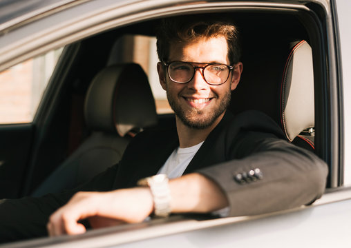 Attractive young man with a beard and glasses smiles while looking out the window of his car.