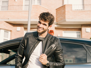 Attractive young man with a beard smiles at camera and leans on his car.