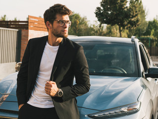 Close-up of attractive young man with beard and glasses in suit is leaning on his car.